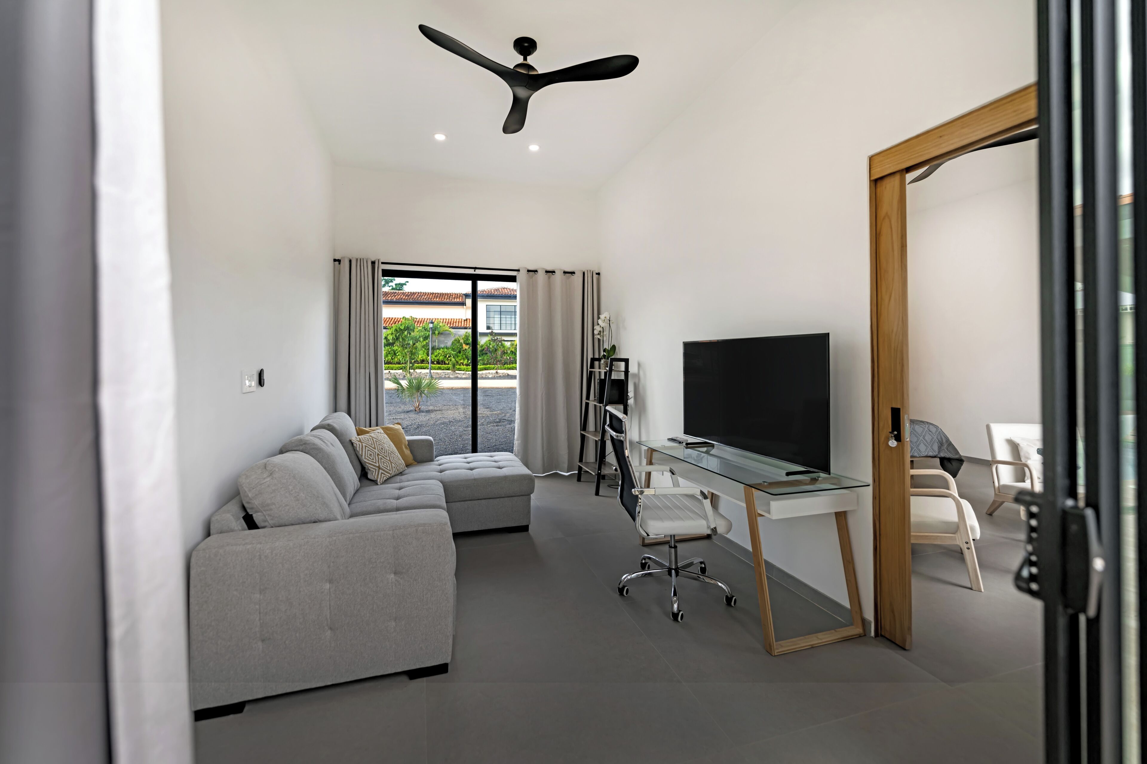 A modern living room with a grey sectional sofa, a glass desk with a flat-screen TV, and a ceiling fan.