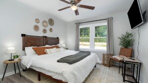 Ein Schlafzimmer mit Bett, Deckenventilator, Fernseher, einem Fenster mit Vorhängen und einer Pflanze.