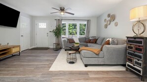 Ein Wohnzimmer mit grauem Sofa, einem Couchtisch, einem an der Wand montierten Fernseher, einem Deckenventilator und einem Fenster mit Vorhängen.