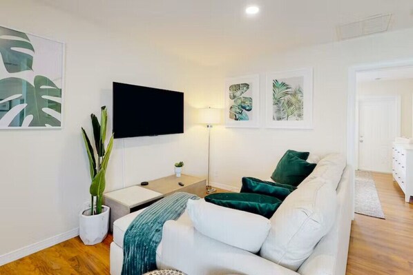 Un salon moderne avec un canapé blanc, des coussins verts, un téléviseur à écran plat, une plante en pot et des œuvres d’art accrochées aux murs.