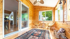 A wooden cabin interior with a large sliding glass door leading to a dining area, two wicker chairs, a patterned rug, and a ceiling fan.