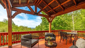 A covered outdoor deck with wooden railings, wicker chairs, a table, and a view of a forested landscape.