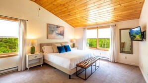 A bedroom with a bed, bedside tables, a bench, a TV, and large windows with curtains.