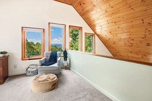 A cozy attic room with a sloped wooden ceiling, large windows offering a view of greenery, a comfortable armchair, and a small round ottoman. - Mountain Views, Hot Tub near Hiking, Ski & Santa`s Village (Jefferson)