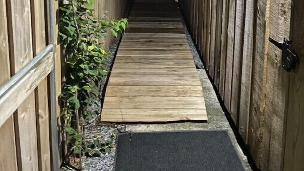 Ein schmaler Holzweg, gesäumt von Holzzaun, führt in einen dunklen Bereich.