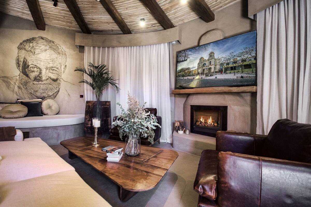 A cozy living room with a fireplace, leather sofas, a coffee table with books and a vase, a large portrait on the wall, and a TV mounted above the fireplace.