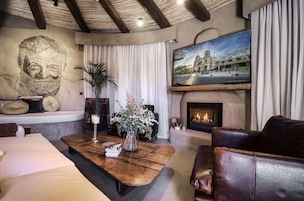 Uma sala de estar acolhedora com lareira, sofás de couro, uma mesa de centro com livros e um vaso, um retrato grande na parede e uma televisão montada acima da lareira.