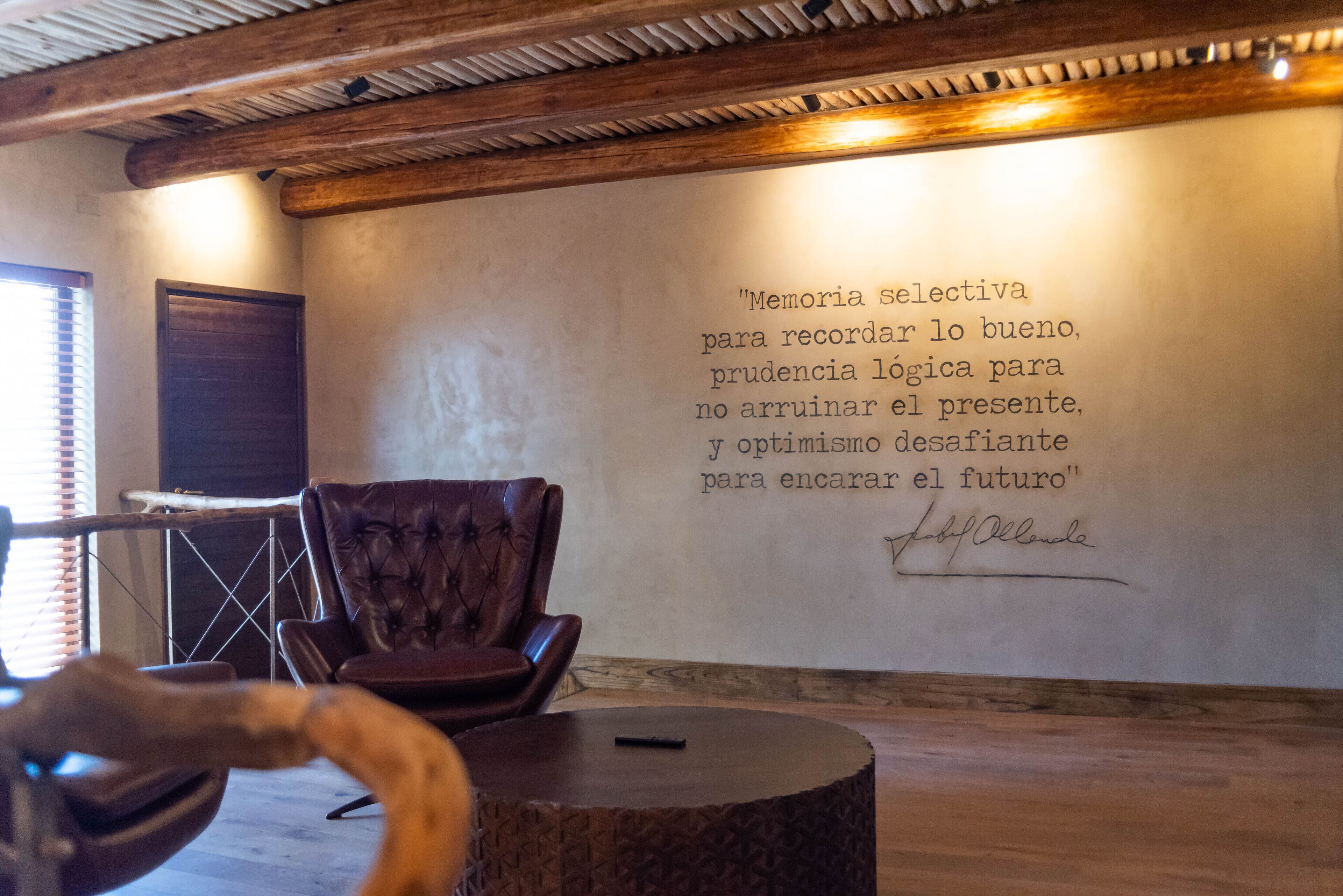 Una habitación con techo de madera, un sillón de cuero, una mesa redonda de madera y una pared con una cita en español.