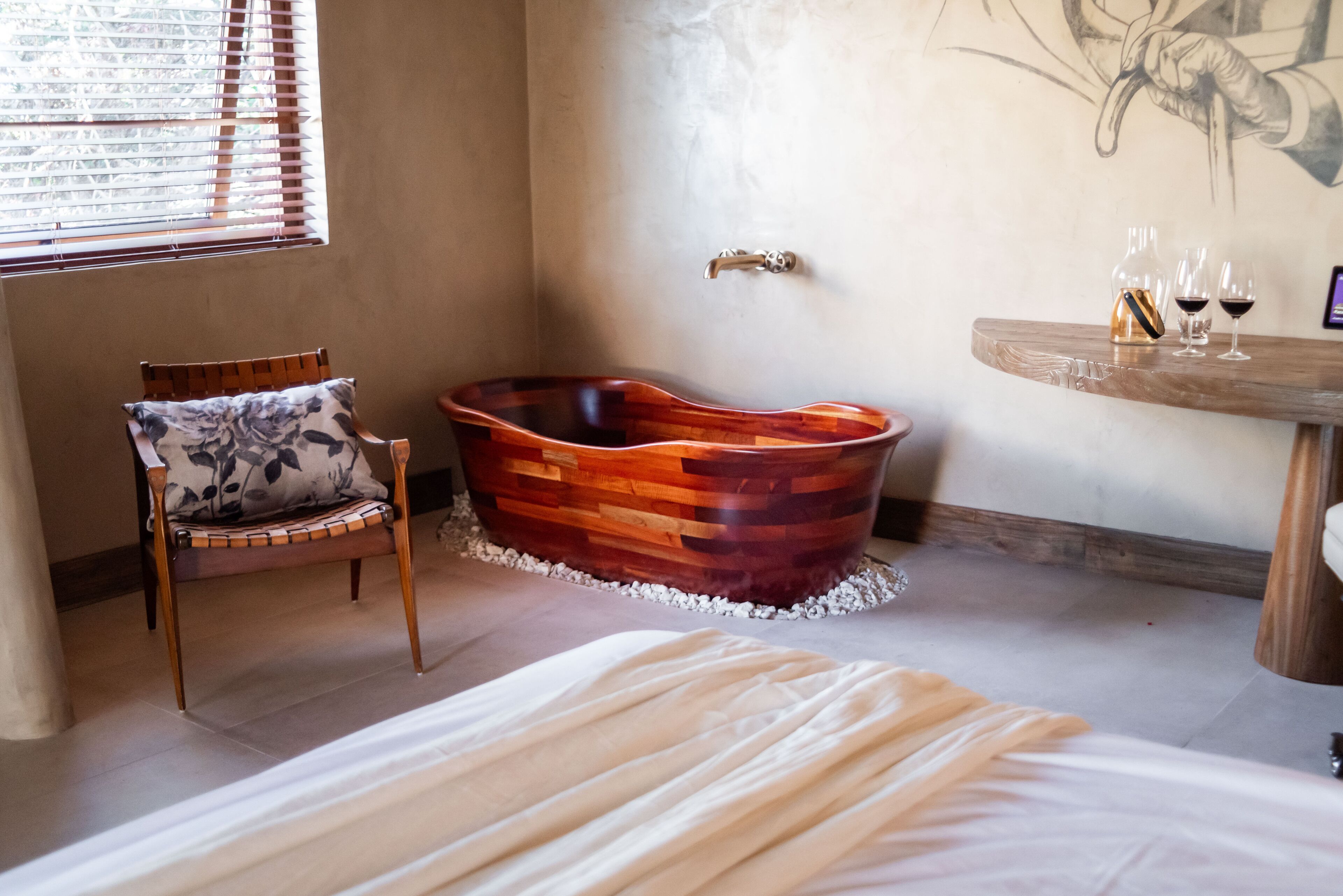 A room with a wooden bathtub, a chair with a floral cushion, a bed with a white bedspread, and a wall-mounted faucet.