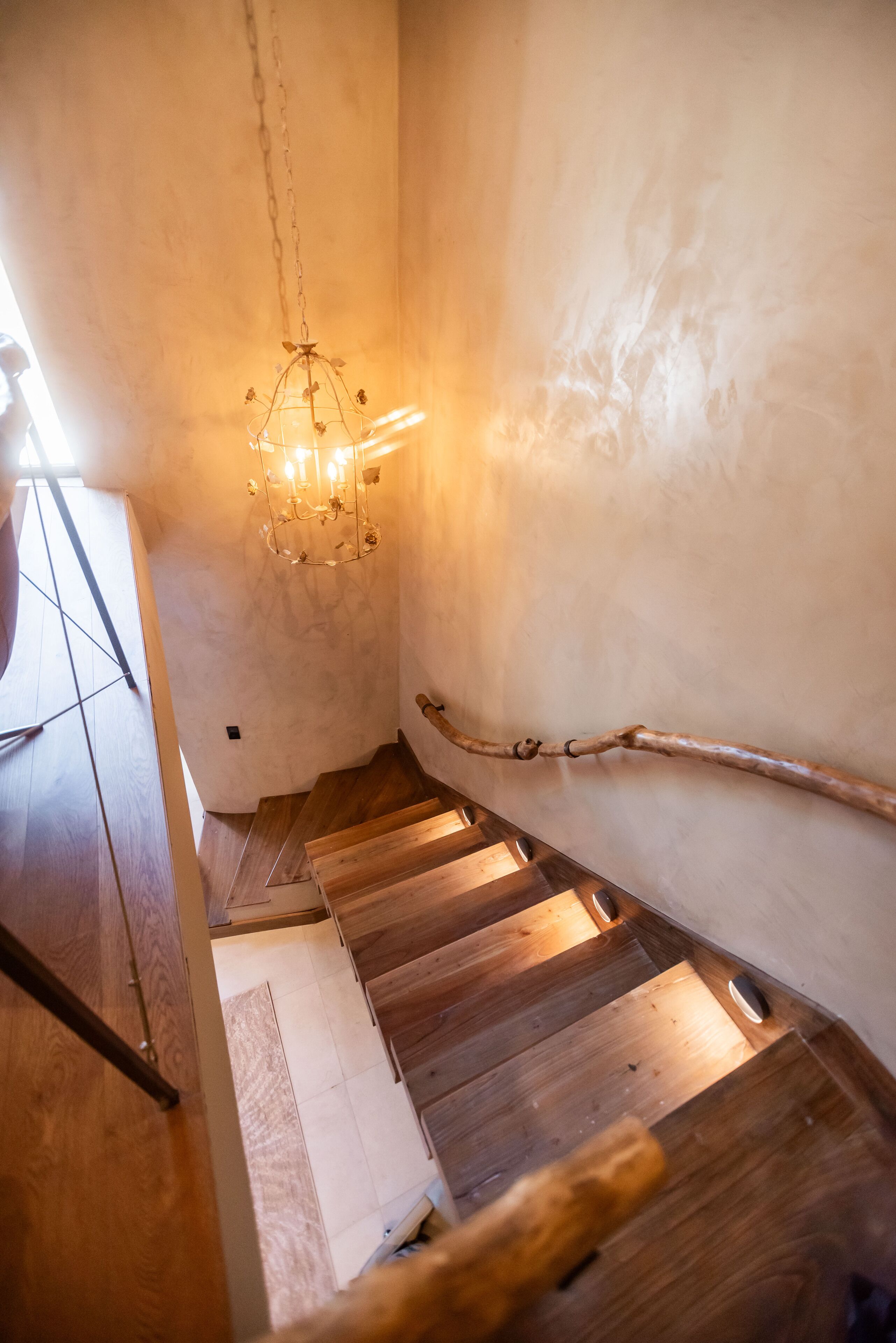 A wooden staircase with a chandelier, a wooden handrail, and a textured wall.