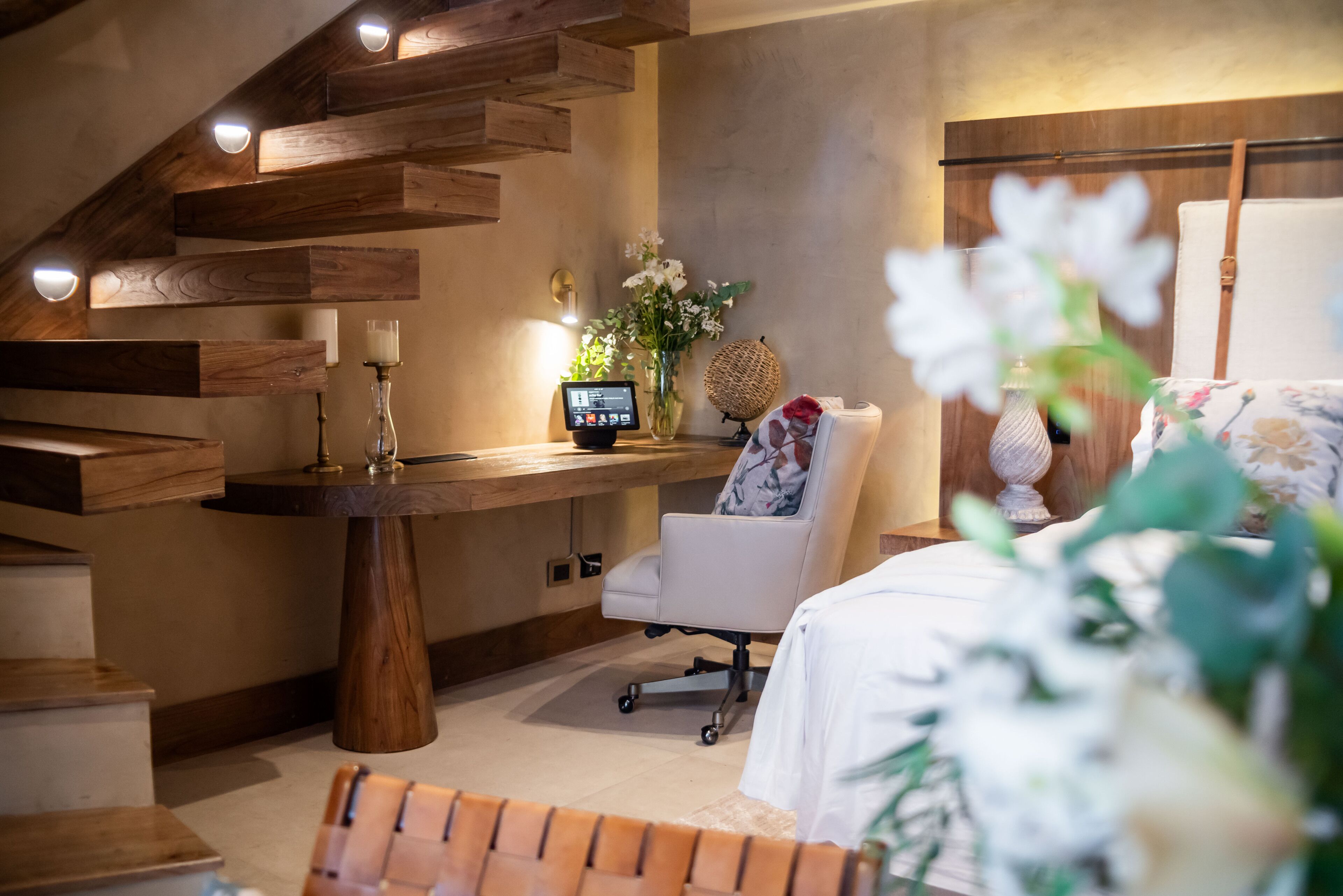 A modern room with a wooden staircase, a desk with a computer, a white chair, and a bed with floral bedding.