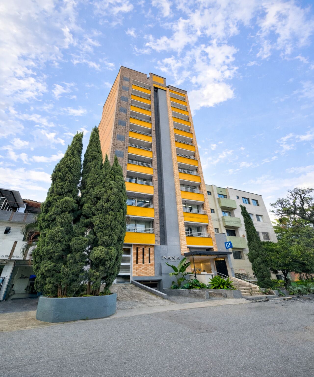 A tall building with yellow and grey exterior, surrounded by greenery and other buildings.