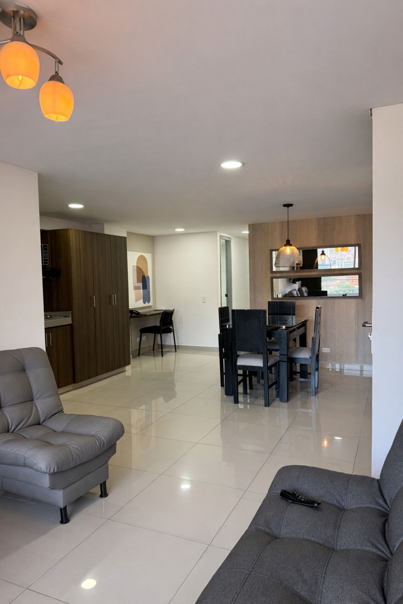 A modern living room with a grey sofa, a dining area with a table and chairs, and a kitchen with wooden cabinets and a window.
