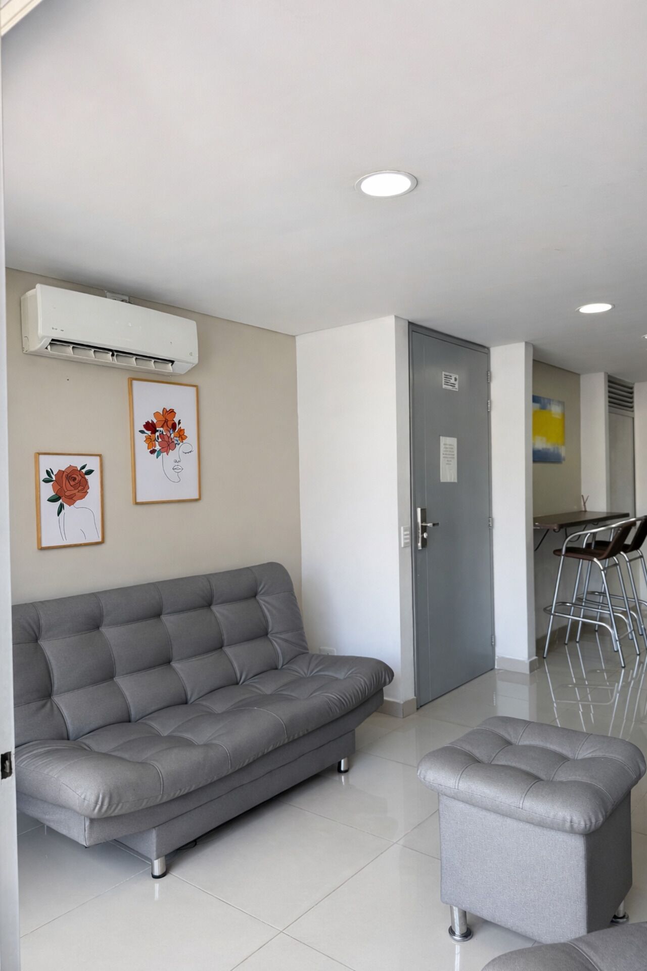 Ein modernes Wohnzimmer mit einem grauen Sofa, einem kleinen Hocker und einem Essbereich mit Metallstühlen.