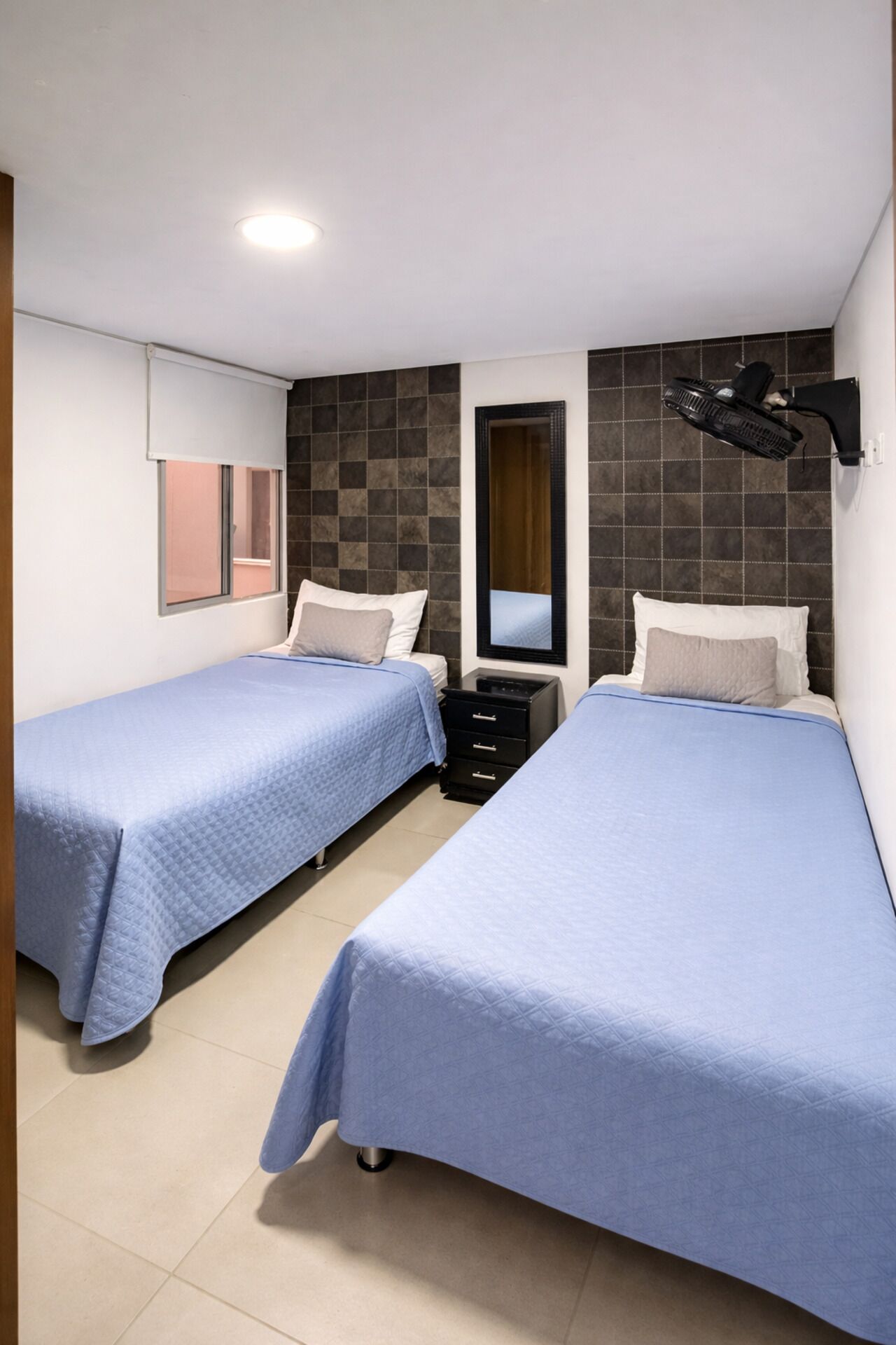 Ein Hotelzimmer mit zwei Einzelbetten, einem kleinen Schminktisch, einem Spiegel und einem Deckenventilator.