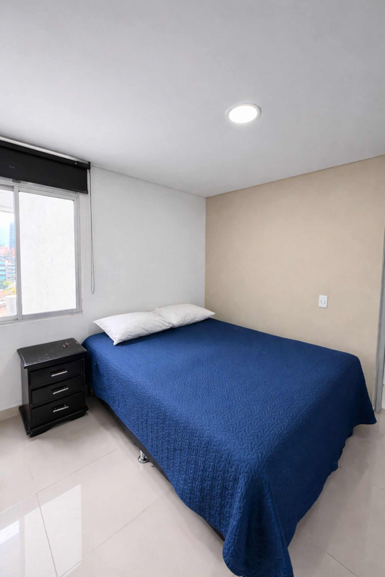 Un dormitorio con cama, mesita de noche, ventana y lámpara de techo.