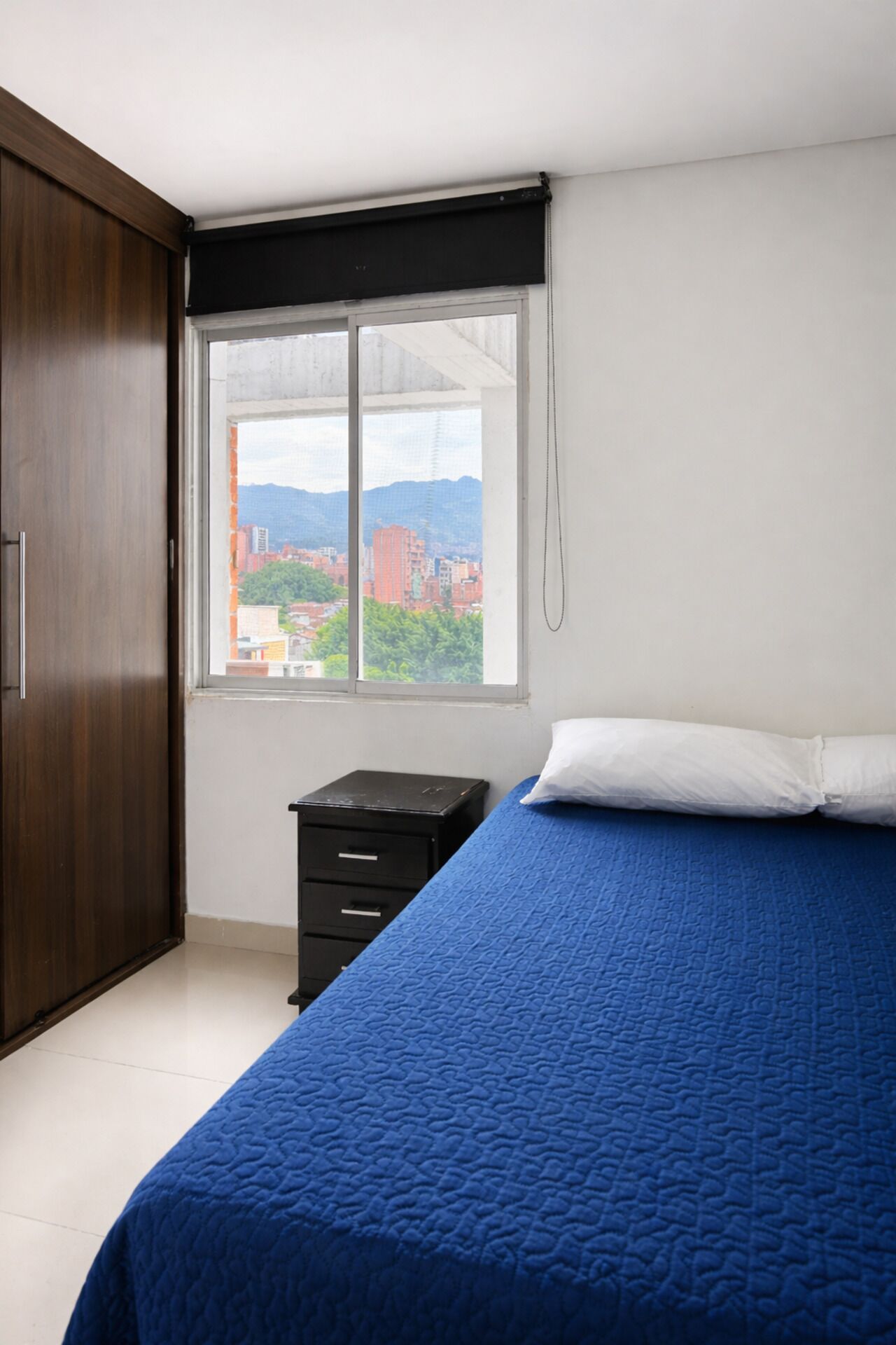 A bedroom with a large window, a bed with a blue bedspread, a wooden wardrobe, and a small black dresser.