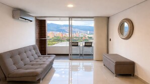 Een ruime woonkamer met een bank, een lange zitbank en een grote schuifpui die uitkomt op een balkon met uitzicht op de stad.