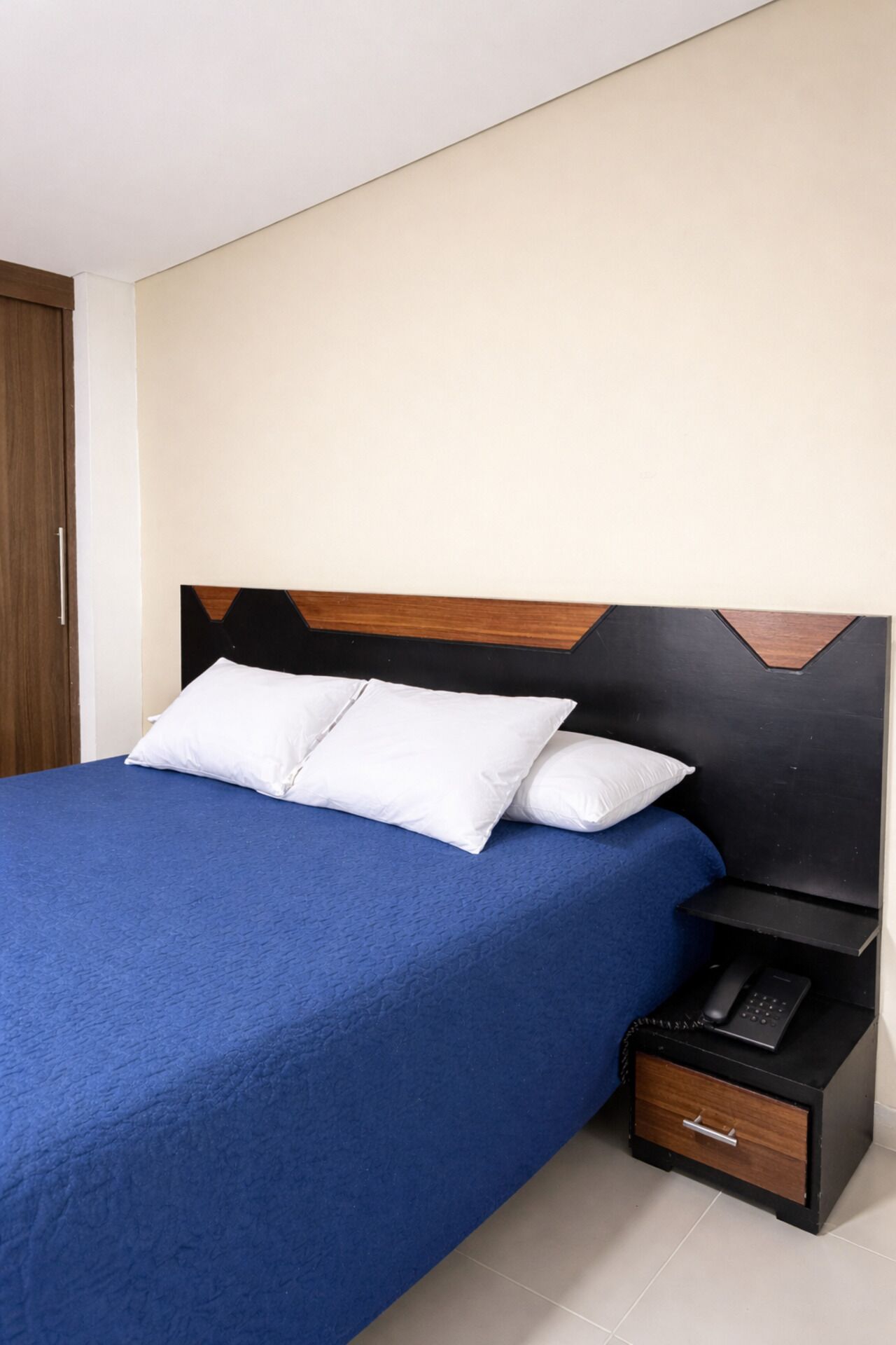 A bed with a blue bedspread, two white pillows, a wooden headboard, and a bedside table with a phone and a drawer.