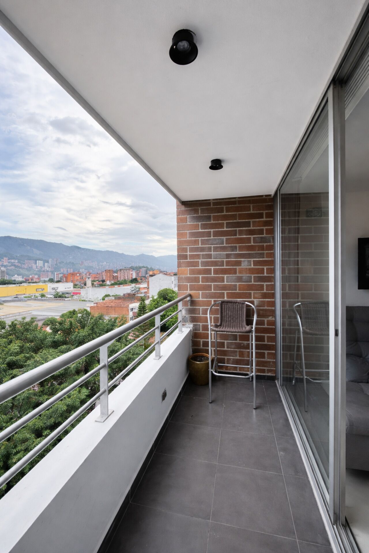 A balcony with a view of a cityscape, a brick wall, and a glass door leading to a room.