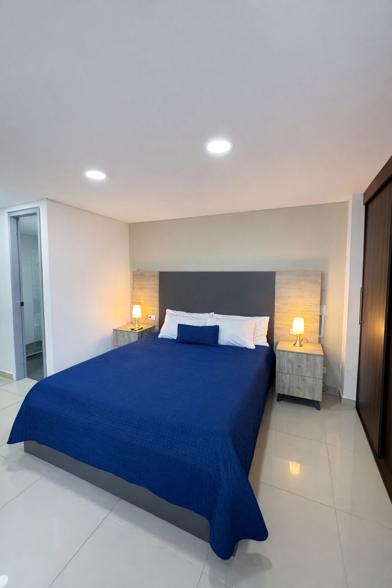 A modern bedroom with a large bed, two bedside tables with lamps, a wooden headboard, and a door to the left.