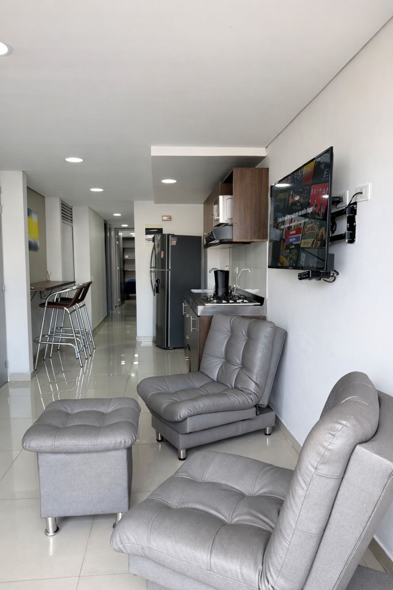 A modern living room with a grey sectional sofa, a small table with chairs, a kitchen area with a refrigerator and stove, and a wall-mounted TV.