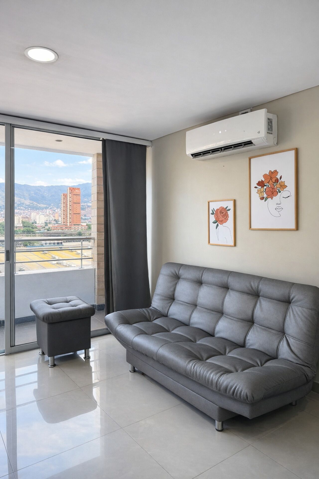 A modern living room with a grey sofa, a small ottoman, and a city view through a large window.