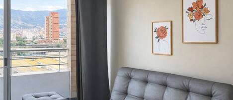 A modern living room with a grey sofa, a small ottoman, and a city view through a large window.