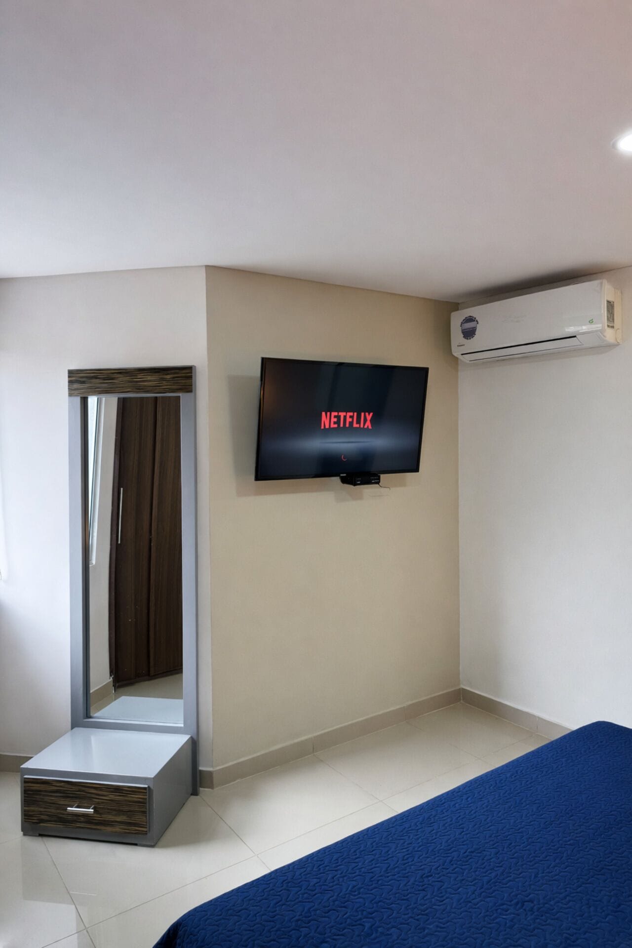A hotel room with a bed, a television displaying 'NETFLIX,' a mirror, and an air conditioning unit.