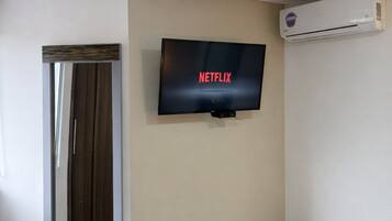 A hotel room with a bed, a television displaying 'NETFLIX,' a mirror, and an air conditioning unit.
