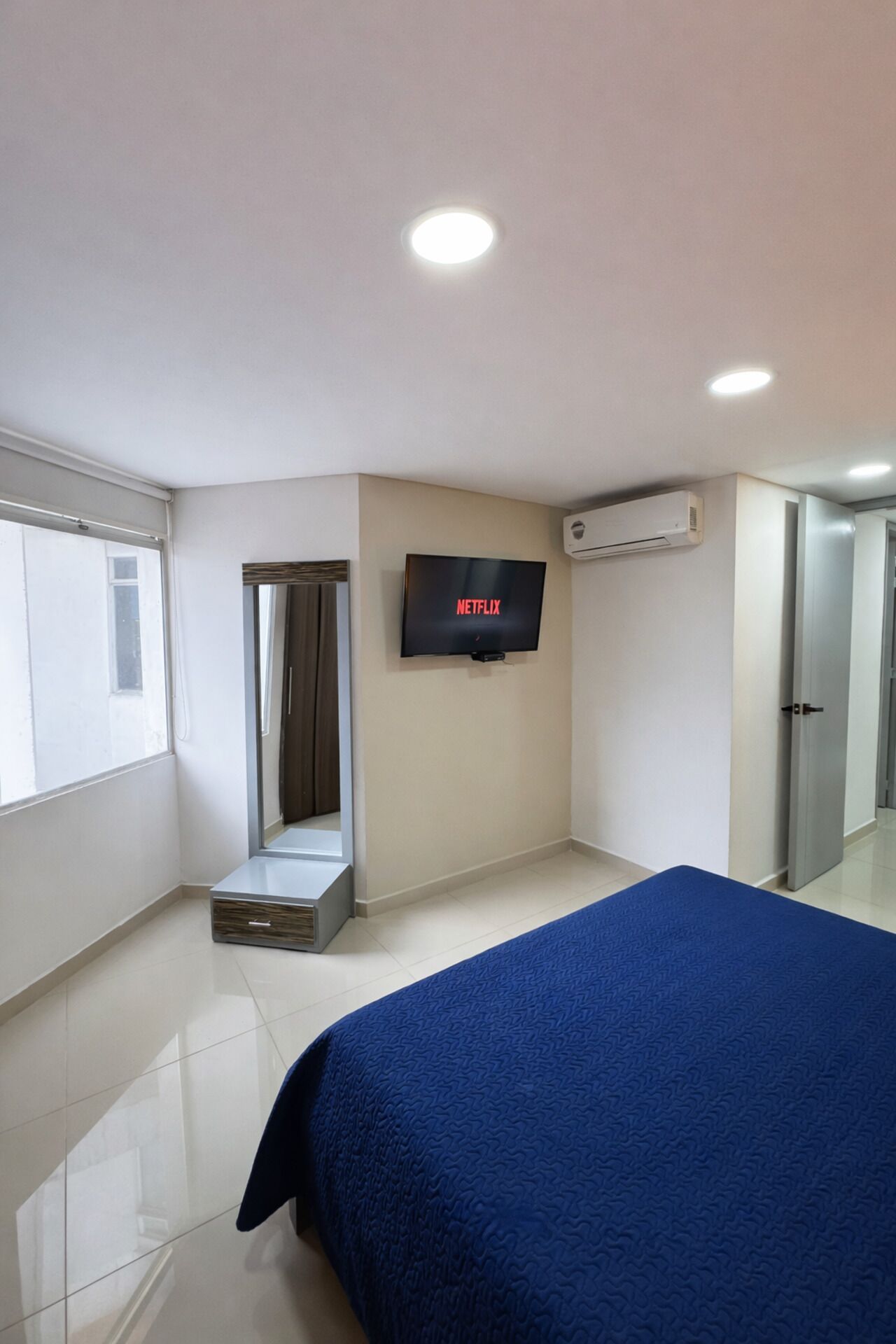 A bedroom with a bed, a TV mounted on the wall, an air conditioning unit, a wardrobe, and a window.