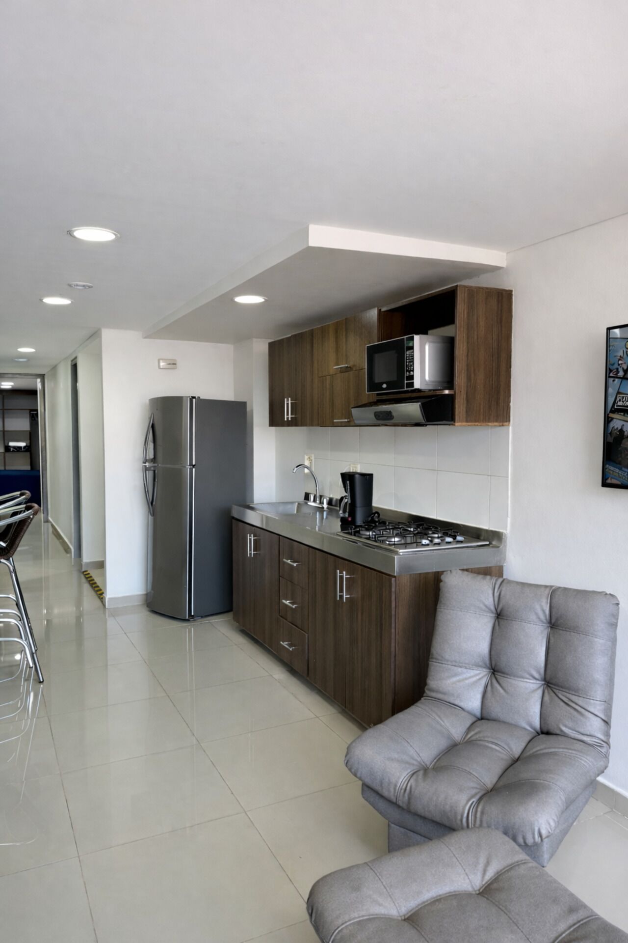 A modern kitchen with a built-in microwave, stove, and refrigerator. There is a grey upholstered armchair and a dining area with a table and chairs.