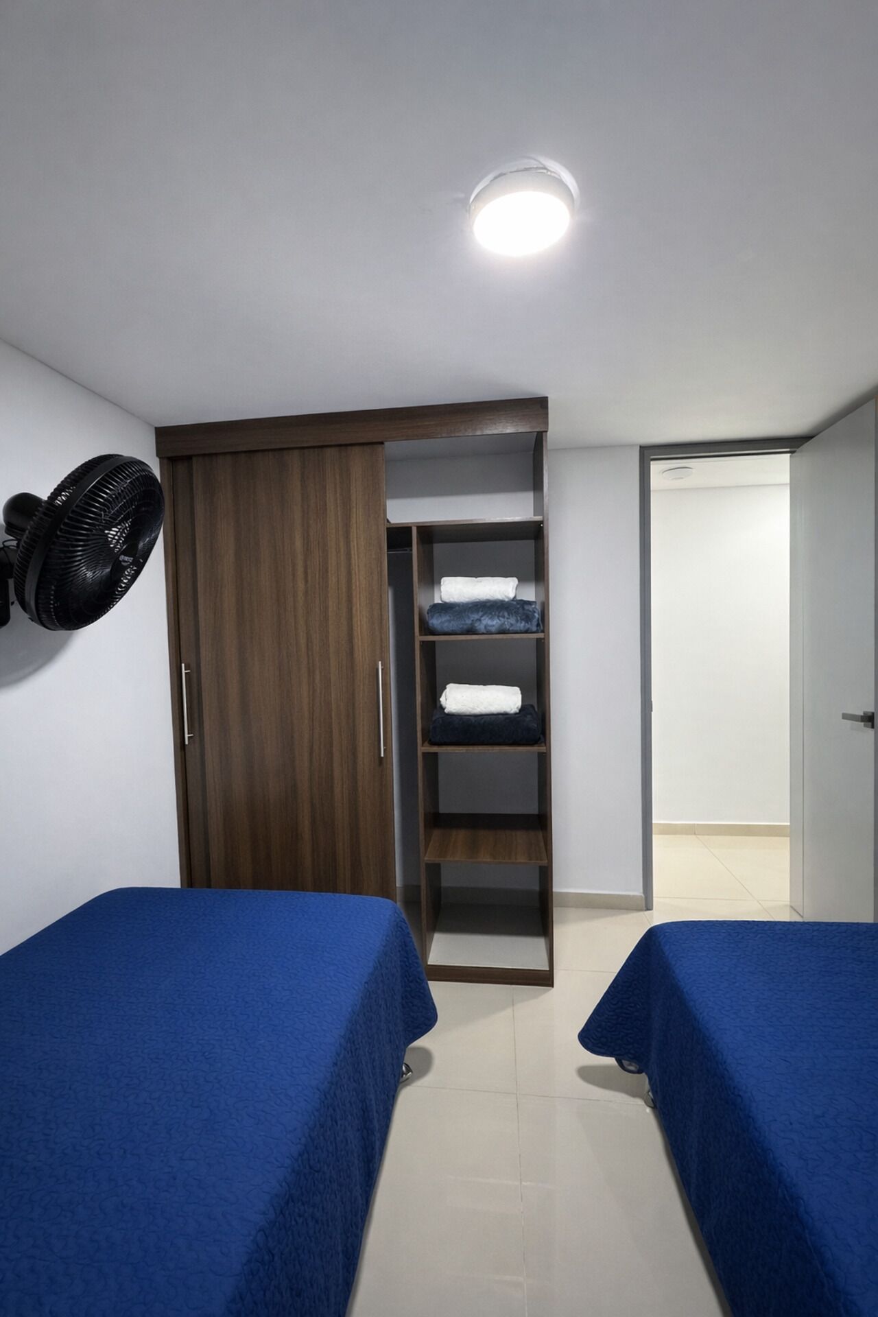 Una habitación con dos camas, un armario, una estantería con ropa doblada y un ventilador.