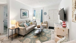 A living room with a sofa, coffee table, and television. There is a door and a window with curtains.