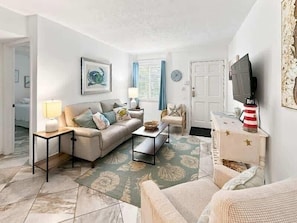 A living room with a sofa, coffee table, and television. There is a door and a window with curtains.