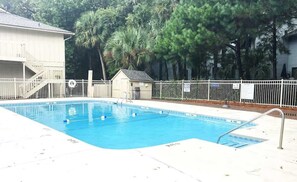 A rectangular swimming pool with a white deck and metal railings, surrounded by a white fence and palm trees.