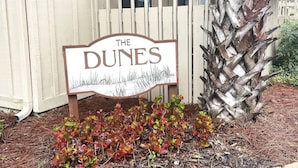 A sign reading "The Dunes" with a wooden fence and a palm tree in the background.