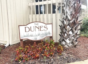 A sign reading "The Dunes" with a wooden fence and a palm tree in the background.