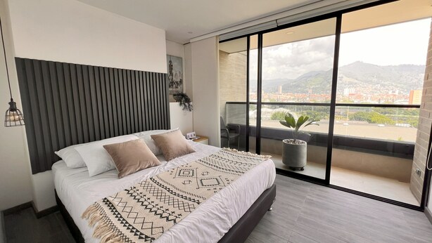 A modern bedroom with a large bed, a view of the city, and a balcony with a plant. - Spacious loft at amazing location! (sabaneta)