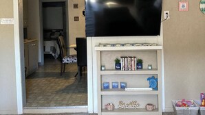 Ein Wohnzimmer mit Deckenventilator, einem Flachbildfernseher an einer weißen Kommode, einem Bücherregal mit Büchern und Dekorationsobjekten sowie einer Tür zu einem anderen Raum.