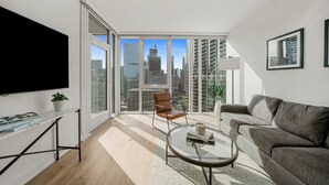 Una sala moderna con un televisor grande, un sofá, una mesa de centro y una silla. Se ve un ventanal con vistas a la ciudad.