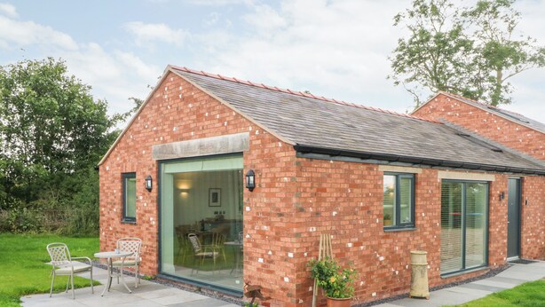 A brick building with a large glass door entrance, a patio with outdoor seating, and a small garden area. - Curlew (Malpas)