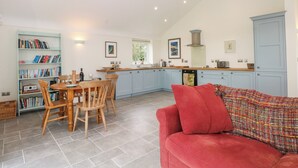 A spacious living area with a red sofa, a wooden dining table with chairs, a bookshelf filled with books, and a kitchen with light blue cabinets and a chimney. - Curlew (Malpas)
