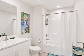 A bathroom with a white sink, toilet, and bathtub. There is a painting on the wall and a wicker basket. - Sea Glass 308 | Gulf Shores, AL (Gulf Shores)