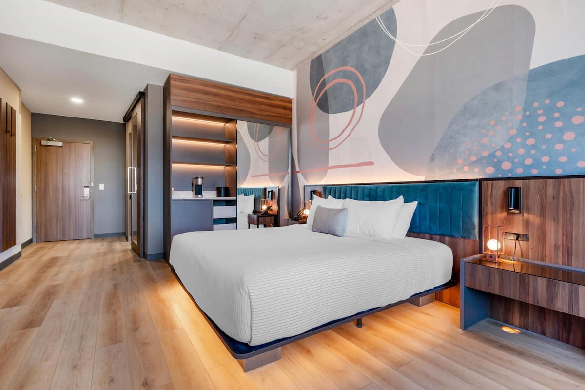 A modern hotel room with a large bed, wooden headboard, and a wall with abstract art.