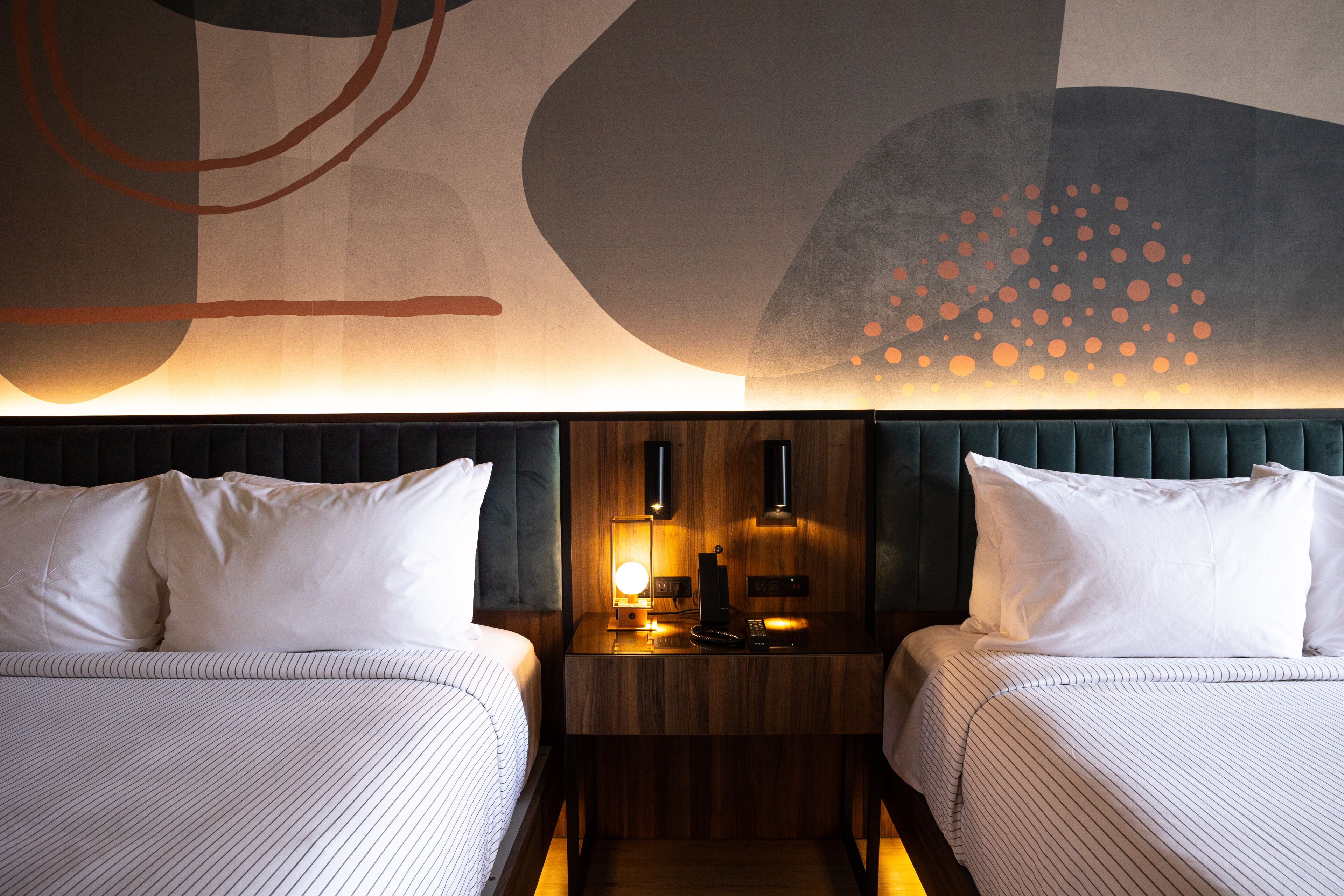 Una habitación de hotel con dos camas, cabecero de madera, mesita de noche con lámpara y arte abstracto en la pared.