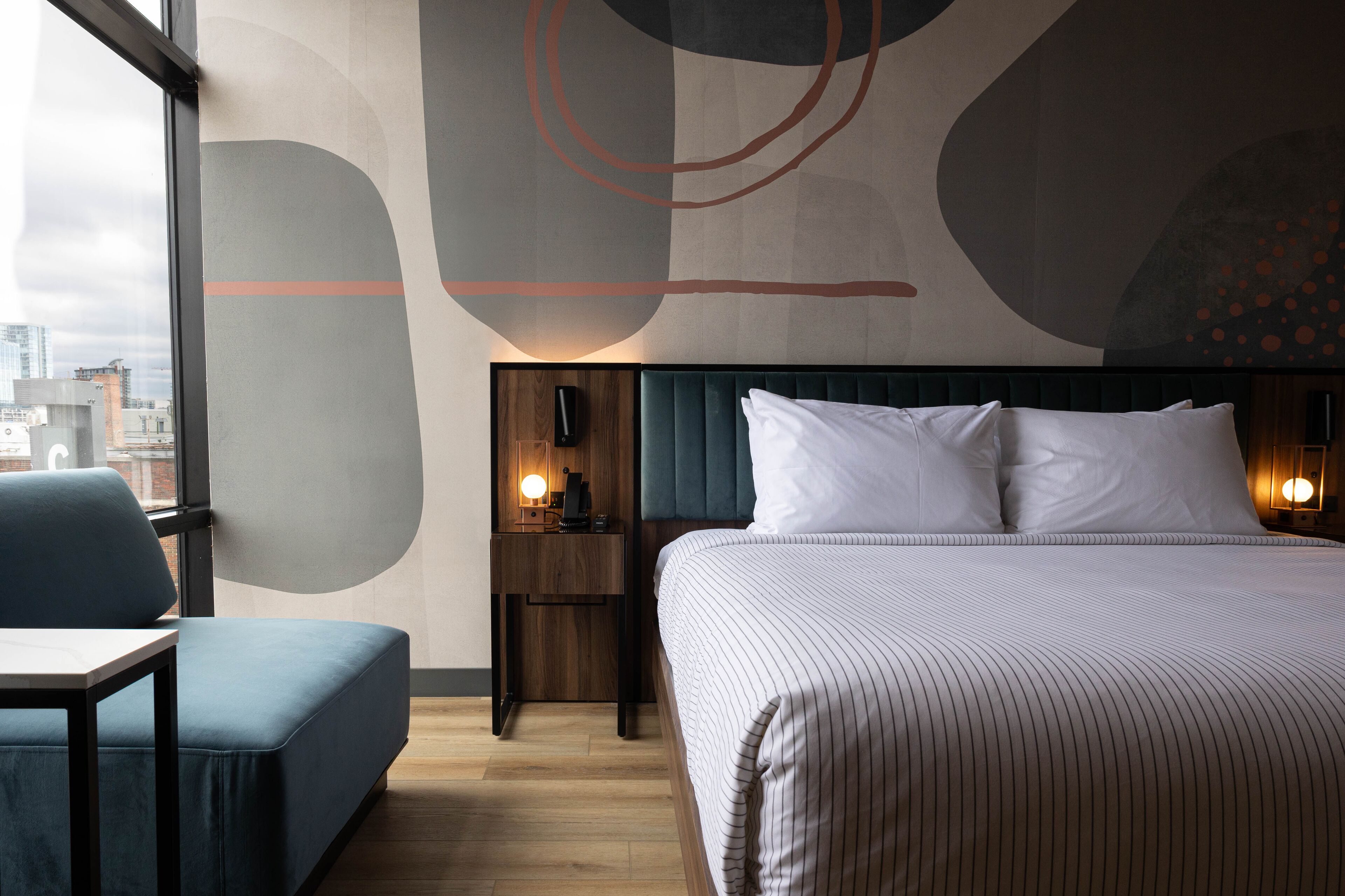 A hotel room with a large bed, a nightstand with a lamp, a seating area with a chair, and a view of the city through large windows.