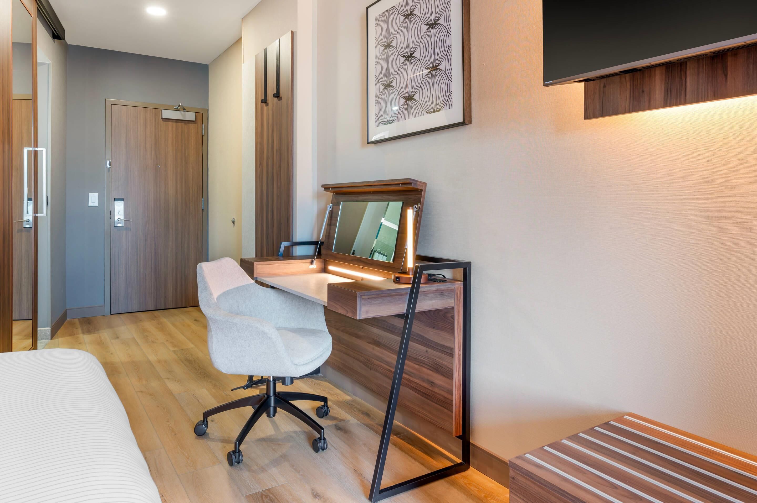 Una moderna camera d'albergo con una scrivania in legno, una sedia da ufficio bianca, un letto e un grande specchio.