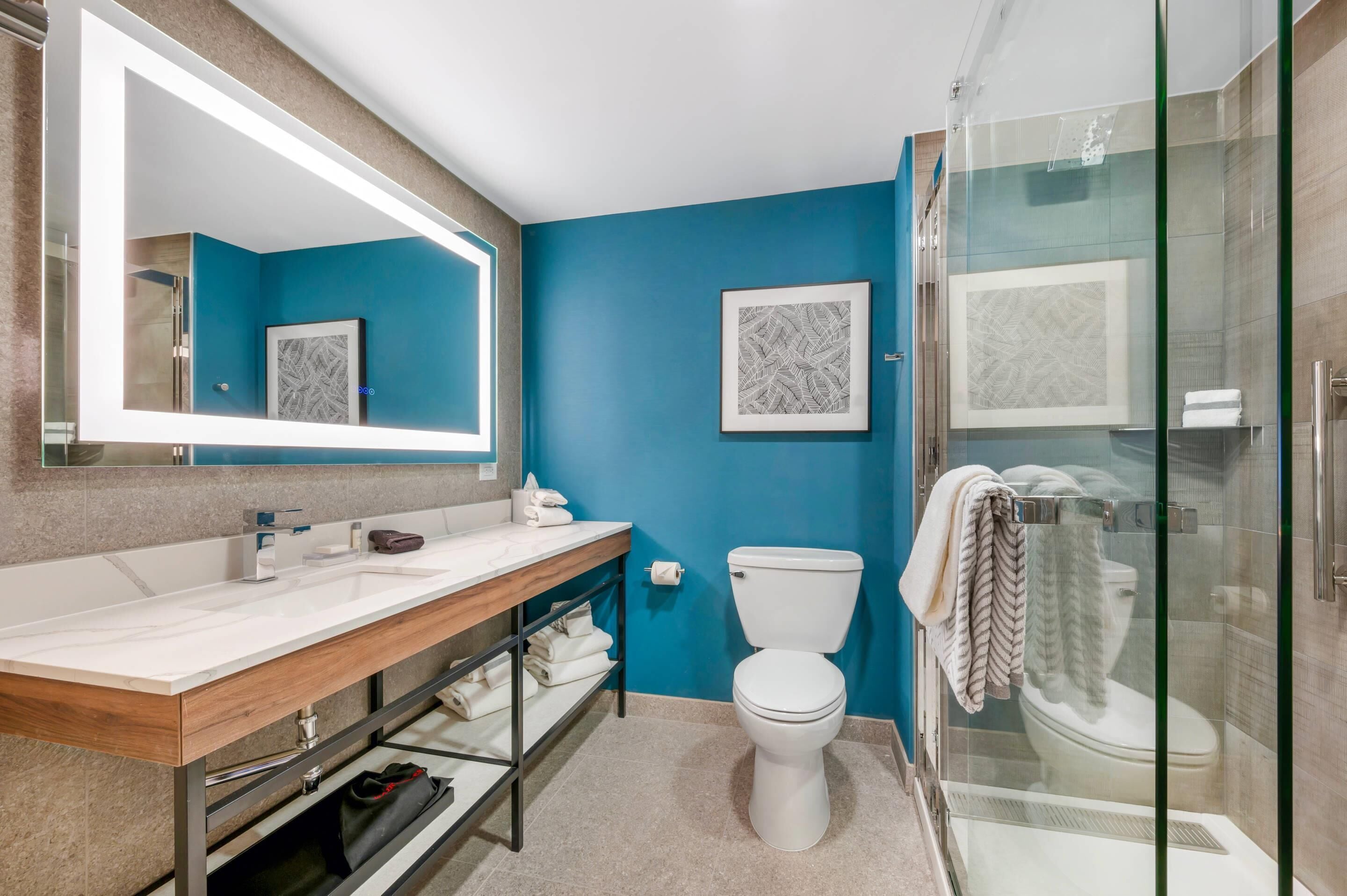 Een badkamer met een blauwe wand, een wit toilet, een douche met glazen wanden en een houten wastafelmeubel met spiegel.