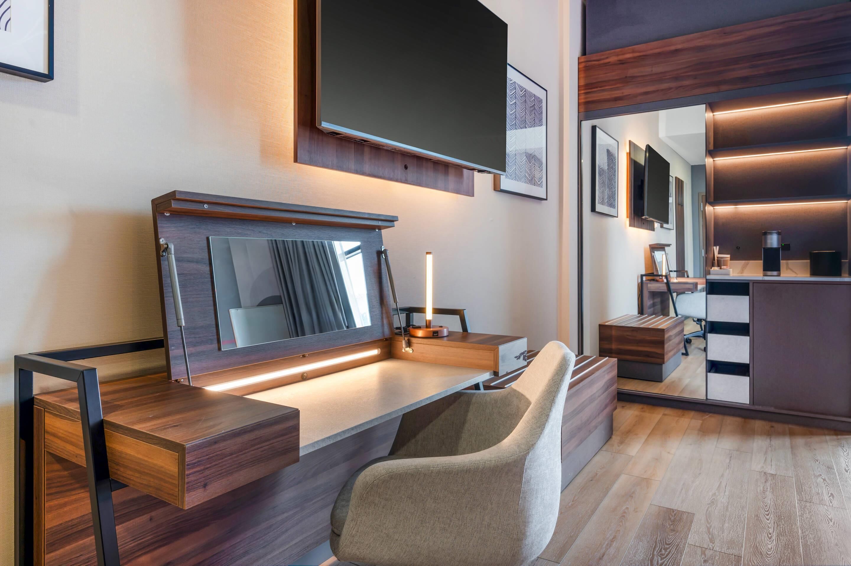 A modern hotel room with a wooden desk, a comfortable chair, a large mirror, and a flat-screen TV mounted on the wall.